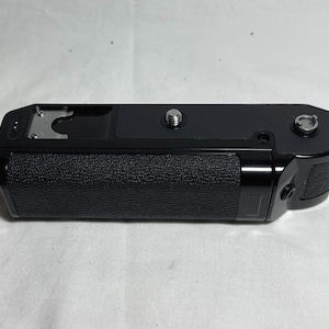 Puede incluir: Empuñadura de cámara negra con superficie texturizada. Cuenta con un tornillo metálico y un botón plateado. La parte superior tiene una placa metálica y pequeños agujeros.