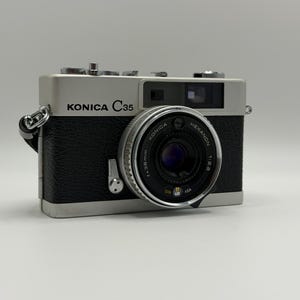 May include: A silver and black Konica C35 rangefinder camera featuring a Konica Hexanon 38mm f/2.8 lens. The front of the camera body displays the text Konica C35, while the lens barrel includes technical markings for aperture and film speed.