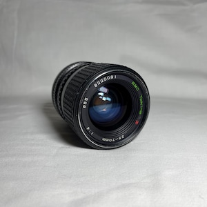 Puede incluir: Un objetivo de cámara RMC Tokina negro de 35-70 mm f/4. El objetivo tiene un anillo de enfoque acanalado y marcas, incluyendo "8220091" y "ø55". El objetivo está ligeramente inclinado hacia la izquierda.