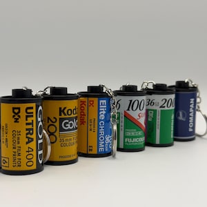 Pode incluir: Uma coleção de chaveiros de rolos de filme. Os rolos exibem marcas como Kodak e Fuji. As cores dos rolos são variadas, incluindo amarelo, azul e verde. Cada chaveiro tem um anel prateado.