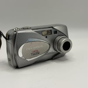 May include: A silver Olympus Camedia C-460 Zoom digital camera featuring a compact rectangular body, a prominent lens barrel on the right, a built-in flash, and a brown wrist strap attached to the left side. The front panel displays the brand name.