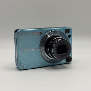 May include: A light blue Sony Cyber-shot digital camera with a metallic brushed finish. The front features a prominent black Carl Zeiss Vario-Tessar lens, a built-in flash, and visible wear marks on the lower left corner of the camera body.