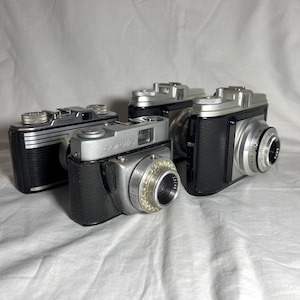 Może przedstawiać: Cztery zabytkowe aparaty fotograficzne z srebrnymi i czarnymi korpusami ułożone na białej powierzchni. Aparaty mają obiektywy i tarcze, a na jednym z nich widnieje napis "Dersityle". Aparaty są w dobrym stanie.