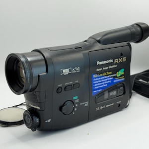 Puede incluir: Una videocámara Panasonic RX5 negra con lente gran angular, zoom de 14x, estabilizador de imagen y compartimento para cinta VHS-C. El panel lateral muestra especificaciones técnicas, incluyendo 0,7 Lux de sensibilidad y ahorro de energía.