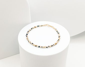 May include: A delicate bracelet featuring small, round beads in cream, black, and brown hues. The beads are strung together with a gold-colored chain and clasp. The bracelet is displayed on a white pedestal against a white background.