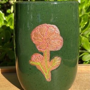 May include: A dark green ceramic mug with a floral design. The flower is a light brown color with pink accents and green stems and leaves. The mug is cylindrical and appears to be handmade.