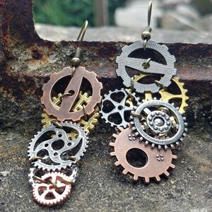 Könnte beinhalten: Steampunk-Ohrringe mit einer Ansammlung von Metallzahnrädern in verschiedenen Größen und Farben. Die Ohrringe zeigen eine Mischung aus bronzefarbenen, silberfarbenen und goldfarbenen Zahnrädern, die einen einzigartigen, industriellen Look erzeugen.