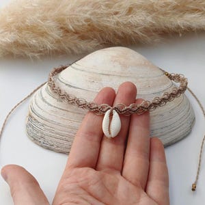 Könnte beinhalten: Eine braune Makramee-Halskette mit einem weißen Muschelanhänger. Die Kette ist um eine Hand gebunden und liegt auf einer Muschel.