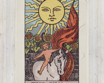 Könnte beinhalten: Eine cremefarbene Decke mit einem Tarotkarten-Design. Die Karte zeigt eine große gelbe Sonne mit einem Gesicht, ein Kind, das ein weißes Pferd reitet, und den Text "THE SUN". Der Hintergrund umfasst Sonnenblumen und ein rotes Banner.