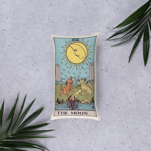 May include: Rectangular pillow featuring a tarot card design. The image depicts a yellow sun with a face, a crescent moon, two dogs, and a crayfish. The text "THE MOON" is at the bottom, with "XVIII" at the top. The background is light blue.