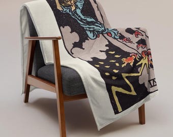 May include: A white throw blanket draped over a gray armchair. The blanket features a tarot card design with a figure in blue robes, a black background, and yellow and red accents. The chair has wooden arms and legs.