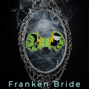 May include: Green and black Frankenstein and Bride shoe wings enamel pins in a silver ornate mirror frame. The pins feature a green monster and a green bride with black hair. The text "Franken Bride Shoe Wings" is below the mirror. The website address "www.TragicStudio804.com" is below the text.