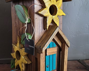 May include: A wooden birdhouse with a teal blue door and two yellow sunflower accents. The birdhouse has a small wooden platform and a brown roof. The birdhouse has a tag that says "Idahoing".