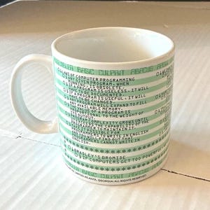 Puede incluir: Una taza de cerámica blanca con rayas horizontales de color verde claro y texto en verde oscuro. El texto impreso enumera varias leyes de la programación informática, incluyendo líneas como 'Any given program, when running, is obsolete'.