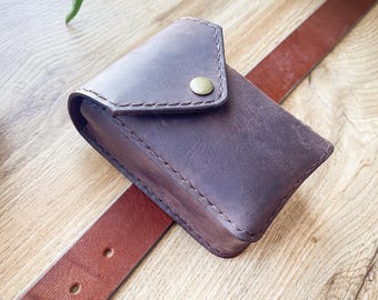 Puede incluir: Una cartera de cuero marrón con cierre de solapa y un botón de latón. La cartera está sobre un cinturón de cuero marrón.
