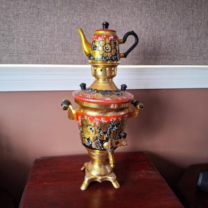 May include: A gold-colored samovar with a matching teapot on top. The samovar and teapot are decorated with floral patterns in red, black, and gold. The samovar has a spigot and handles. The background is a brown wall and a wooden table.