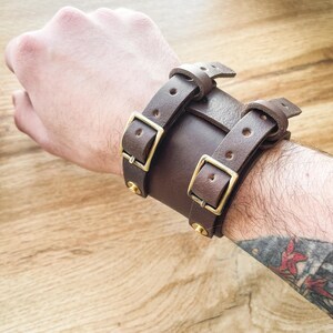 Può includere: Un braccialetto in pelle marrone con due cinturini regolabili e fibbie in ottone. Il braccialetto ha borchie decorative in ottone.