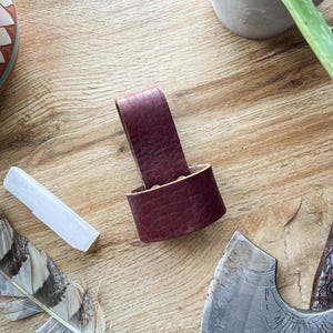 Puede incluir: Un lazo de cinturón de hacha de cuero burdeos, diseñado para sujetar un hacha. El lazo está hecho de cuero grueso y texturizado. Otros elementos en la imagen incluyen una pluma, un cristal, una maceta decorativa y un hacha pequeña.