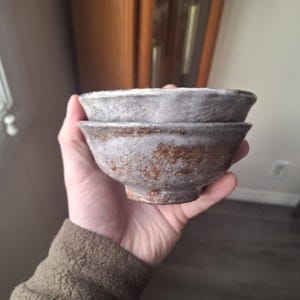 May include: Two handmade ceramic bowls stacked on top of each other. The bowls have a rustic, textured appearance with a mix of brown, gray, and white tones. The bowls are held in a hand, showcasing their size and shape.