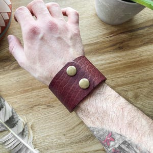 Peut inclure: Un bracelet de manchette en cuir bordeaux avec deux fermoirs bouton en laiton.