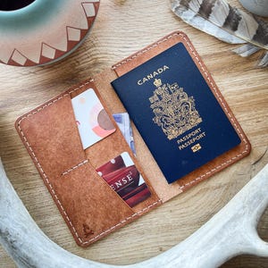 Puede incluir: Un portapasaporte de cuero marrón con un pasaporte canadiense en su interior. El portapasaporte tiene dos ranuras para tarjetas y un bolsillo para dinero en efectivo. El portapasaporte está cosido con hilo blanco.