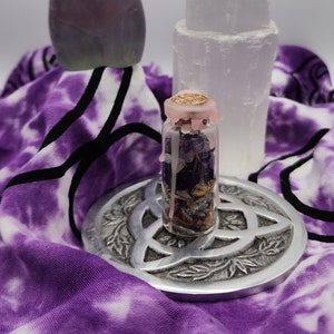 Puede incluir: Una pequeña botella de vidrio llena de cristales, hierbas y flores secas, sellada con una tapa de cera rosa. La botella está sentada sobre un diseño de triqueta de metal plateado, rodeada de una tela de teñido anudado púrpura y blanca. Un gran cristal transparente y un cristal morado están en el fondo.