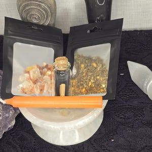 Puede incluir: Un conjunto de tres artículos para un ritual o hechizo. El conjunto incluye una pequeña botella de vidrio con un tapón de corcho, una pequeña bolsa de hierbas secas y una pequeña bolsa de cristales naranjas. Los artículos están dispuestos sobre una superficie blanca.