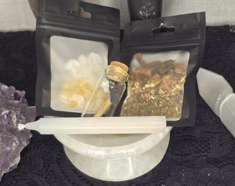 May include: Two small black zippered pouches with clear windows, one containing a small glass bottle with a cork stopper and clear crystals, the other containing dried herbs and spices. A white candle and a white ceramic bowl are also visible.