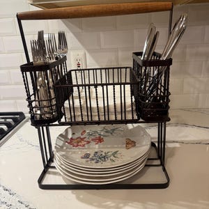 May include: A two-tiered black metal caddy with a wooden handle. The top level features three wire compartments holding silver forks, knives, and a stack of white napkins. The bottom level holds a stack of white plates with floral and butterfly patterns.