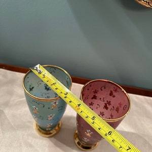 K&ouml;nnte beinhalten: Zwei kleine Gl&auml;ser mit goldenen R&auml;ndern und Blumenmustern. Das Glas links ist hellblau mit wei&szlig;en Blumen und das Glas rechts ist burgunderrot mit rosa Blumen. Ein gelbes Ma&szlig;band ist teilweise im Bild zu sehen.