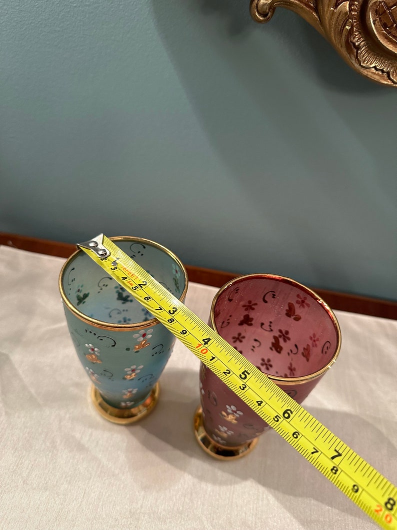 K&ouml;nnte beinhalten: Zwei kleine Gl&auml;ser mit goldenen R&auml;ndern und Blumenmustern. Das Glas links ist hellblau mit wei&szlig;en Blumen und das Glas rechts ist burgunderrot mit rosa Blumen. Ein gelbes Ma&szlig;band ist teilweise im Bild zu sehen.
