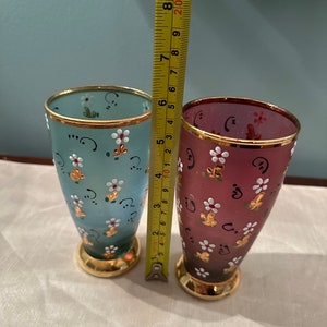 K&ouml;nnte beinhalten: Zwei handbemalte Trinkgl&auml;ser aus Glas mit goldenen Verzierungen. Das Glas links ist hellblau mit wei&szlig;en Blumen und goldenen Akzenten. Das Glas rechts ist dunkelrot mit wei&szlig;en Blumen und goldenen Akzenten.