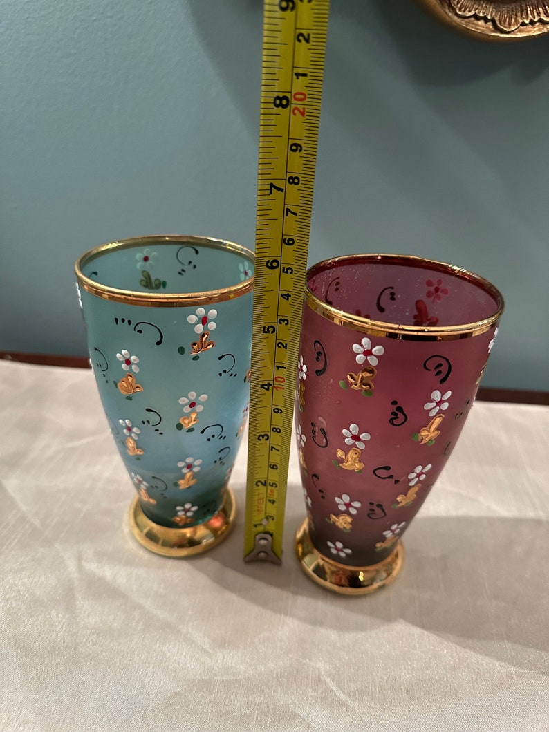 K&ouml;nnte beinhalten: Zwei handbemalte Trinkgl&auml;ser aus Glas mit goldenen Verzierungen. Das Glas links ist hellblau mit wei&szlig;en Blumen und goldenen Akzenten. Das Glas rechts ist dunkelrot mit wei&szlig;en Blumen und goldenen Akzenten.