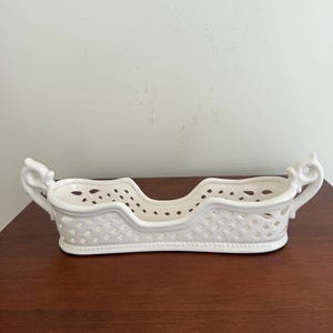 May include: A white ceramic basket with a decorative openwork design. The basket has a rectangular shape with curved sides and two ornate handles. The basket is sitting on a brown surface.