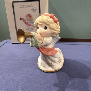May include: A porcelain figurine of an angel with blonde hair and a pink bow, playing a trumpet. The angel is wearing a white dress with a red ribbon and is standing on a base with snowflakes. The base also has the year 2013 inscribed on it. The figurine is part of the Precious Moments collection and is titled "Share the Gift of Love".