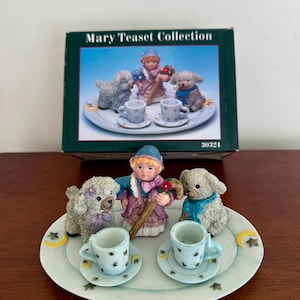 May include: A decorative ceramic teaset from the Mary Teaset Collection. The set includes a figurine of a girl with two dog figurines, two teacups, and saucers. The teacups and saucers are light blue with a floral pattern. The box is green with the collection name.