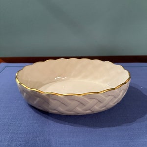 Può includere: Una ciotola in ceramica bianca con un bordo dorato e un motivo intrecciato. La ciotola è su una superficie blu.