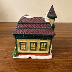 May include: A yellow and green ceramic building with a burgundy roof and white snow. The building has three windows and a small tree in front. The building is part of a Christmas village set.