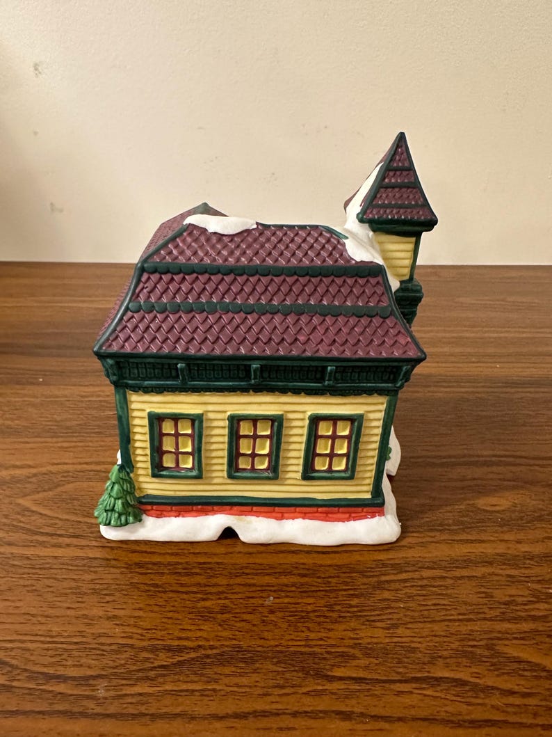 May include: A yellow and green ceramic building with a burgundy roof and white snow. The building has three windows and a small tree in front. The building is part of a Christmas village set.