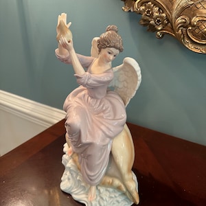 May include: A porcelain figurine of an angel with wings, wearing a pink dress and holding a seashell. The angel is sitting on a seashell with white waves.