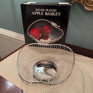 Może przedstawiać: Srebrny koszyk na jabłka w kształcie serca. Koszyk jest pusty i stoi na drewnianym stole. Pudełko na koszyk jest widoczne w tle i zawiera napis "Silver Plated Apple Basket".