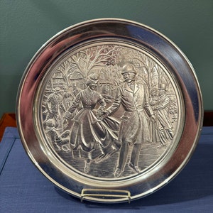 Può includere: Un piatto rotondo argentato con un design in rilievo di una coppia che pattina sul ghiaccio in una scena invernale. Il piatto ha un bordo argentato.