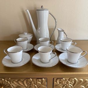 May include: A white porcelain coffee set with silver trim. The set includes a coffee pot, creamer, sugar bowl, and six cups with saucers. The items are arranged on a gold-coloured surface.