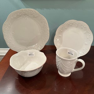 May include: White ceramic dinnerware set with a delicate embossed design. The set includes a large dinner plate, a smaller salad plate, a bowl, and a mug.