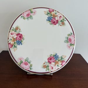 Puede incluir: Un plato redondo de cerámica blanca con un diseño floral. El plato presenta racimos de flores rosas, rojas y azules con hojas verdes alrededor del borde. Un borde rojo oscuro rodea el borde. El plato se exhibe sobre un soporte metálico.