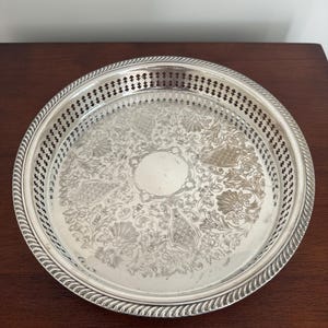 Puede incluir: Una bandeja redonda plateada con un diseño floral grabado y ornamentado. La bandeja tiene un borde perforado y un borde de cuerda trenzada. La bandeja está sobre una superficie de madera marrón oscuro.