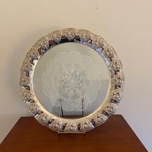 Op de afbeelding: Een zilverkleurige, ronde serveerschaal met een decoratieve bloemenrand. Het midden van de schaal heeft een wit, kantachtig ontwerp. De schaal wordt gepresenteerd op een houten oppervlak.