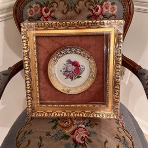 May include: A framed porcelain plate with a gold rim and a floral design featuring pink, white, and purple flowers. The plate is set against a burgundy velvet background.