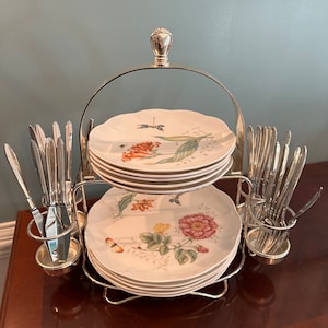 May include: A tiered serving set with floral patterned plates and silverware. The plates have a scalloped edge and feature colorful flowers and butterflies. The set includes a silver metal stand and holders for the silverware.