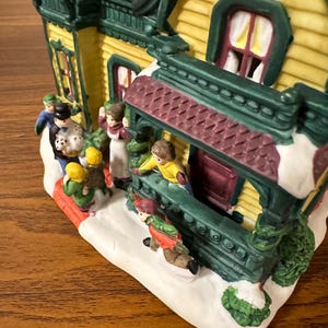 May include: A ceramic Christmas village house with a yellow facade, green trim, and a red roof. The house has a porch with a burgundy railing and a green door. There are small figures of people in front of the house, including a family of four and a couple.